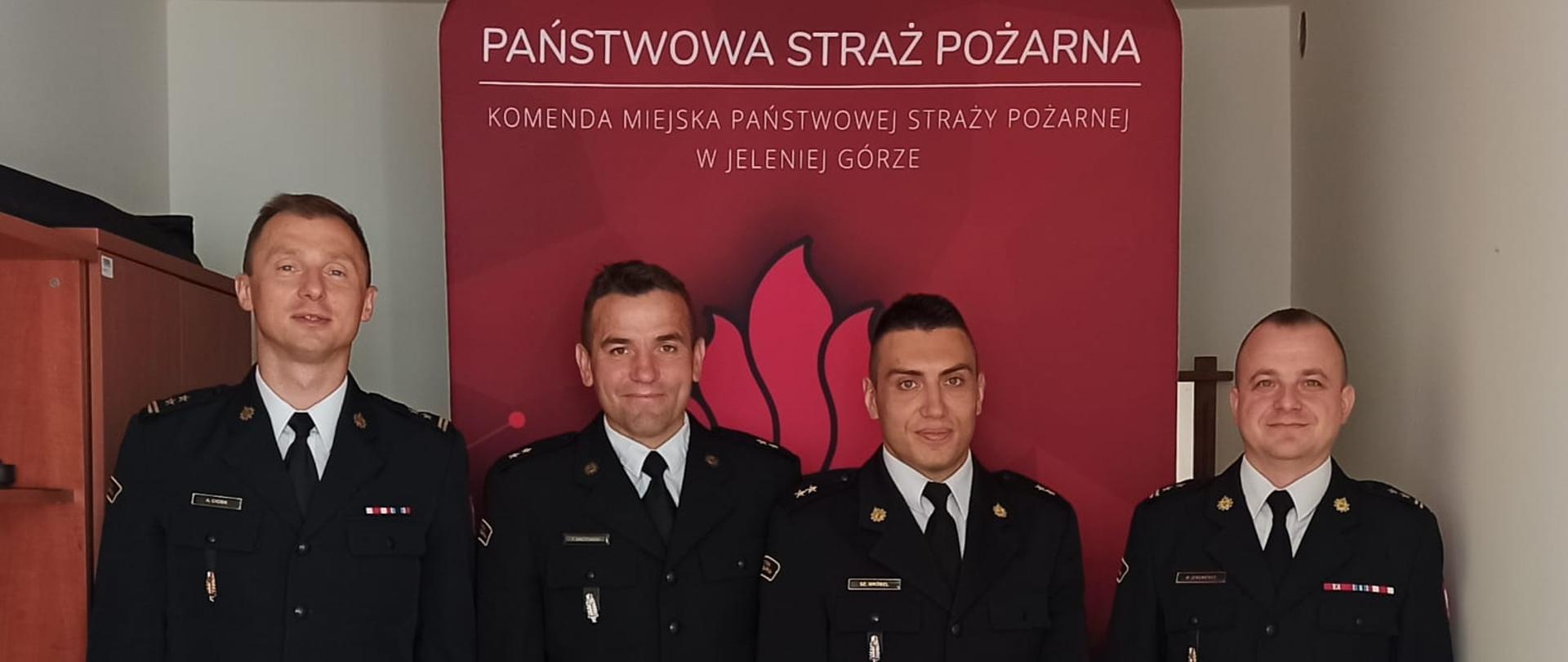 Na zdjęciu widać torsy strażaków w umundurowaniu galowym. Strażacy stoją przed czerwonym banerem z napisem Komenda Miejska Państwowej Straży Pożarnej w Jeleniej Górze w pomieszczeniu biurowym. 