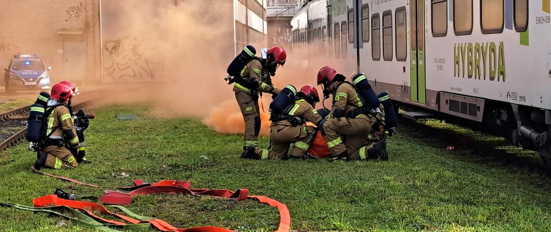 Grupa strażaków w pełnym umundurowaniu prowadzi działania ratownicze przy stojącym na torach pociągu hybrydowym. W tle widoczny jest intensywny dym, a wokół strażaków leżą rozciągnięte węże gaśnicze. Po lewej stronie stoi radiowóz policji.