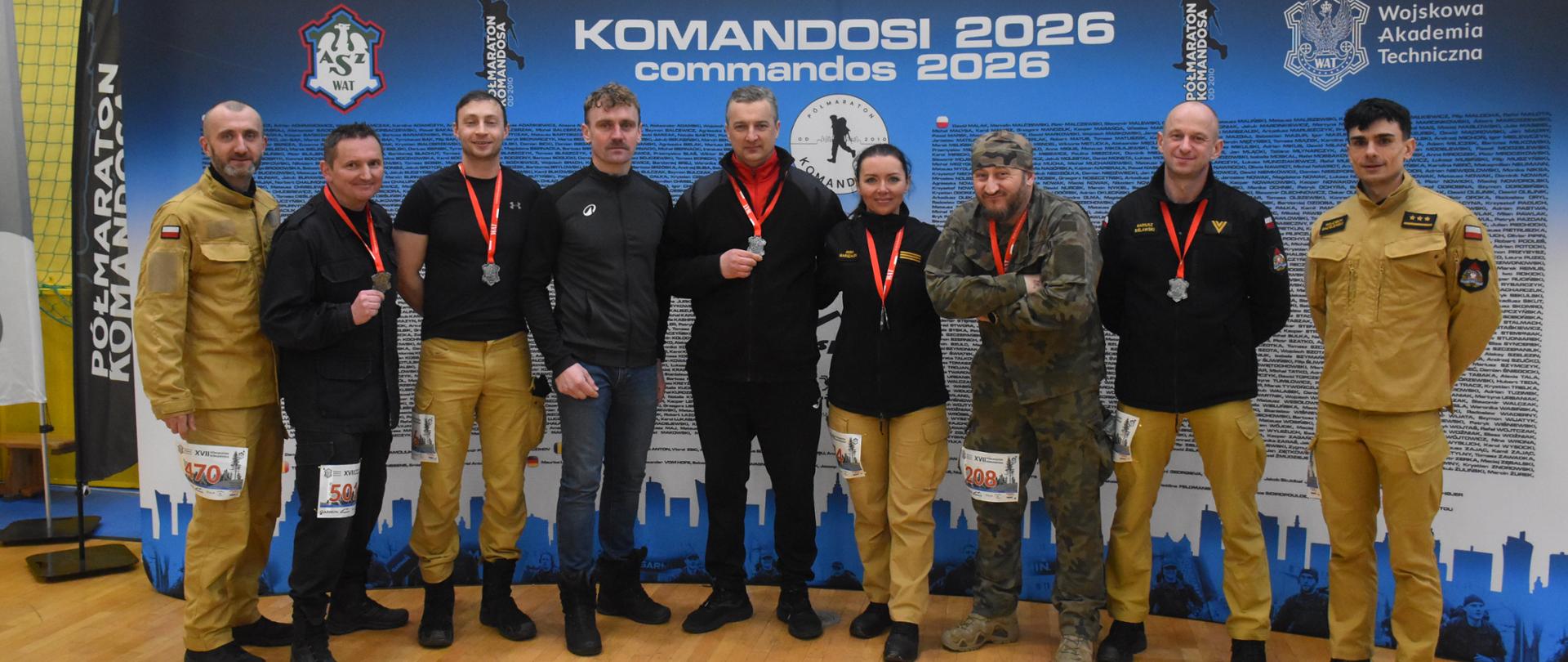 Zdjęcie grupowe uczestników – funkcjonariuszy PSP. Zdjęcie zrobione na Sali sportowej. Uczestnicy stoją przed niebieską ścianką z białymi napisami, m.in. „KOMANDOSI 2026”. 