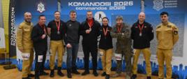 Zdjęcie grupowe uczestników – funkcjonariuszy PSP. Zdjęcie zrobione na Sali sportowej. Uczestnicy stoją przed niebieską ścianką z białymi napisami, m.in. „KOMANDOSI 2026”. 