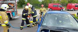 Na pierwszym planie widać czterech druhów z OSP, którzy ćwiczą uwalnianie osoby poszkodowanej
z samochodu osobowego przy użyciu sprzętu hydraulicznego. Dwóch strażaków z Państwowej Straży Pożarnej nadzoruje wykonywanie ćwiczenia. W tle widać 3 samochody pożarnicze.
