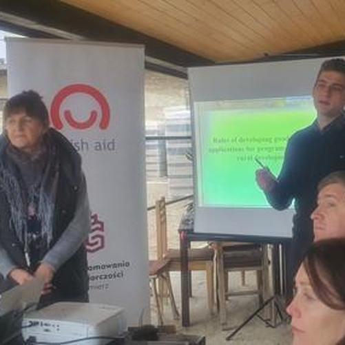 Grupa czterech osób uczestniczy w szkoleniu, w tle rollup z logo Polskiej pomocy oraz ekran i rzutnik multimedialny.