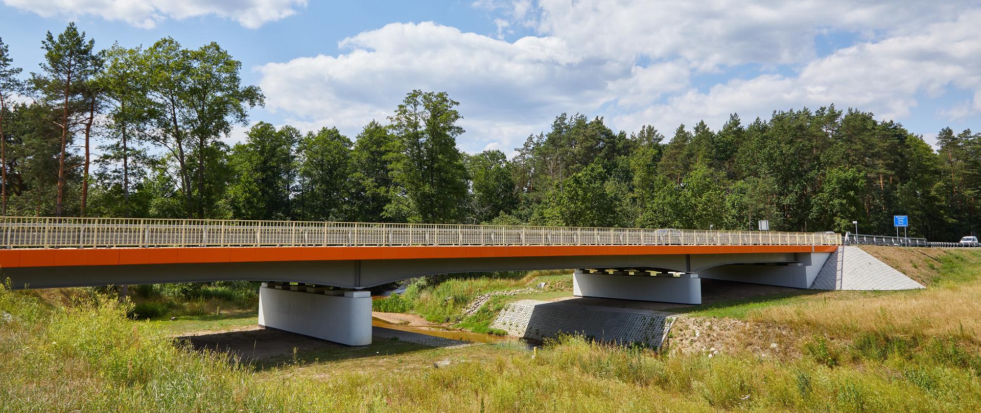 Widok z poziomu ziemi z boku na most. Widać dwie podpory, most wymalowany na kolor pomarańczowy, na moście są barierki. Dookoła łąki. Świeci słońce, trawa zielona. Za mostem widać las i wysokie drzewa.