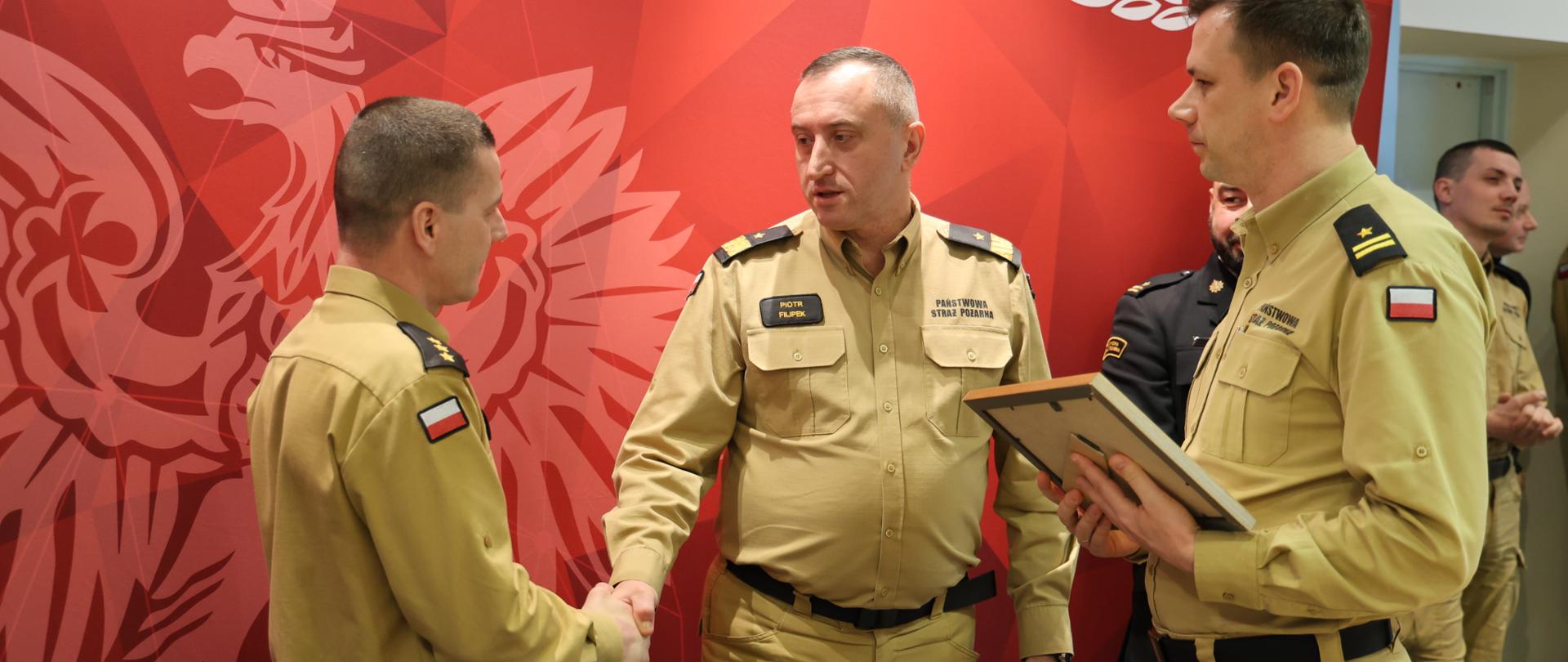 Wnętrze Sali konferencyjnej, na tle czerwonej ścianki z logo Szkoły strażacy w umundurowaniu służbowym i wyjściowym