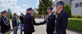 Zdjęcie przedstawia komendanta wojewódzkiego składającego gratulacje prezesowi OSP tytułem wcielenia jednostki do KSRG. Podają sobie prawe dłonie w uścisku, obok po prawej stoi druh osp w umundurowaniu galowym ze sznurem w rogatywce, po lewej stronie komendanta wojewódzkiego stoi wiceprezes zarządu oddziału powiatowego Związku OSP RP w Łowiczu oraz dziennikarze lokalnych mediów, w tle budynek komendy powiatowej.