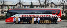 Pamiątkowe zdjęcie grupowe uczestników akcji honorowego krwiodawstwa wraz z pielęgniarkami na tle specjalnego autobusu do pobierania krwi, w tle budynek koszarowy Centralnej Szkoły PSP