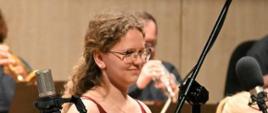 Zdjęcie przedstawiające skrzypaczkę Annę Domską na scenie filharmonii. W tle orkiestra symfoniczna. 