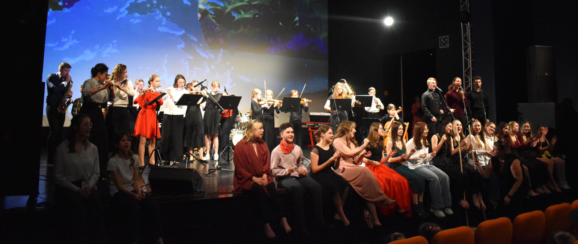 Zdjęcie przedstawia grupę młodych muzyków i wokalistów występujących na scenie. W tle widać kolorową, animowaną projekcję z motywem roślinnym. Na scenie znajdują się osoby grające na instrumentach, takich jak skrzypce, flet, saksofon i inne, oraz grupa osób siedzących z przodu, prawdopodobnie chór lub widownia aktywnie uczestnicząca w wydarzeniu. Scena jest oświetlona reflektorami.