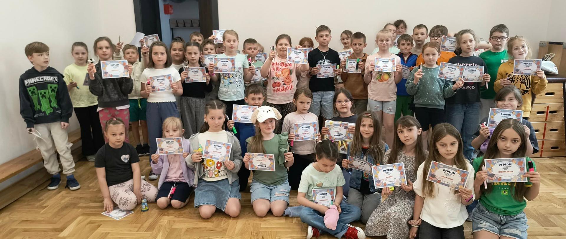 Zdjęcie przedstawia dużą grupę dzieci w wieku wczesnoszkolnym, pozujących do wspólnej fotografii w jasnej sali o białych ścianach i klasycznym, drewnianym parkiecie układanym w jodełkę.
Wszystkie dzieci są ubrane w codzienne, kolorowe stroje letnie – t-shirty, bluzy, spódniczki i krótkie spodenki. Liczna grupa dzieli się na dwa rzędy. Większość dzieci stoi z tyłu, natomiast około dziesięcioro z nich siedzi lub klęczy w pierwszym rzędzie bezpośrednio na podłodze. Niemal każde dziecko dumnie trzyma w dłoniach mały, poziomy, kolorowy dyplom uznania oraz drobne upominki (takie jak długopisy lub flamastry). Na twarzach dzieci malują się uśmiechy oraz skupienie.