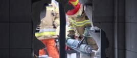 Widok z wnętrza pomieszczenia na strażaka operującego ciężką wiertarką na zewnątrz budynku próbującego powiększyć wykonane przebicie w ścianie