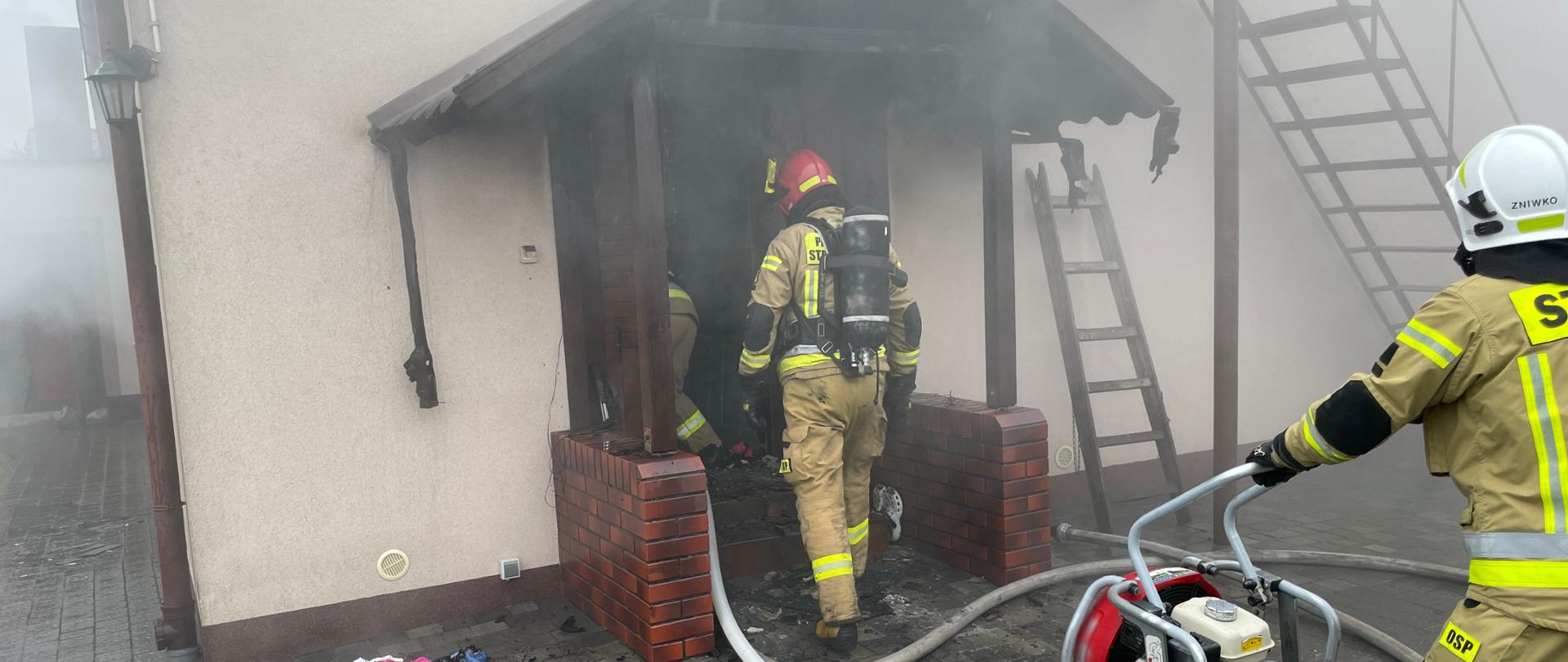 jeden strażak wyposażony w sprzęt OUO wchodzi do budynku przed nim stoi wentylator oraz strażak go obsługujący pod nogami linia wężowa widoczne zadymienie