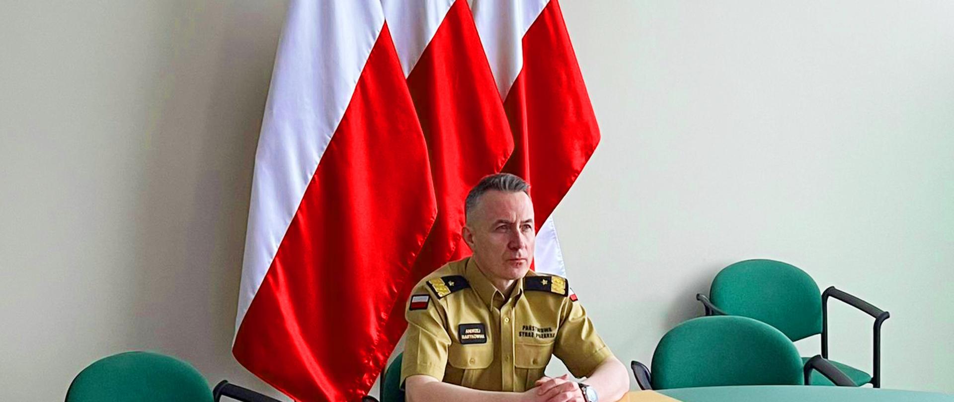 Komendant Główny Państowej Straży Pożarnej nadbryg. Andrzej Bartkowiak w czasie wideokonferencji dotyczącej działań w walce z trzecią falą pandemii COVID-19. W tle flagi narodowe oraz godło 