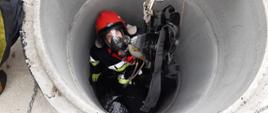 Zdjęcie przedstawia strażaka opuszczonego do studni, ma założone szelki ratownicze, hełm z maską na głowie i rękawice. Na lince ratunkowej ma podczepiony aparat powietrzny.