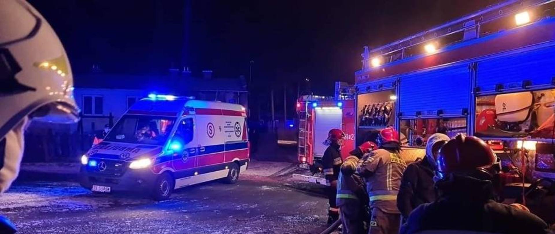 Na zdjęciu widać strażaków chwilę po akcji gaszenia pożaru rozbierających się z aparatów ochrony układu oddechowego, stojących przy samochodzie pożarniczym. W tle widać karetkę pogotowia ratunkowego na sygnałach świetlnych