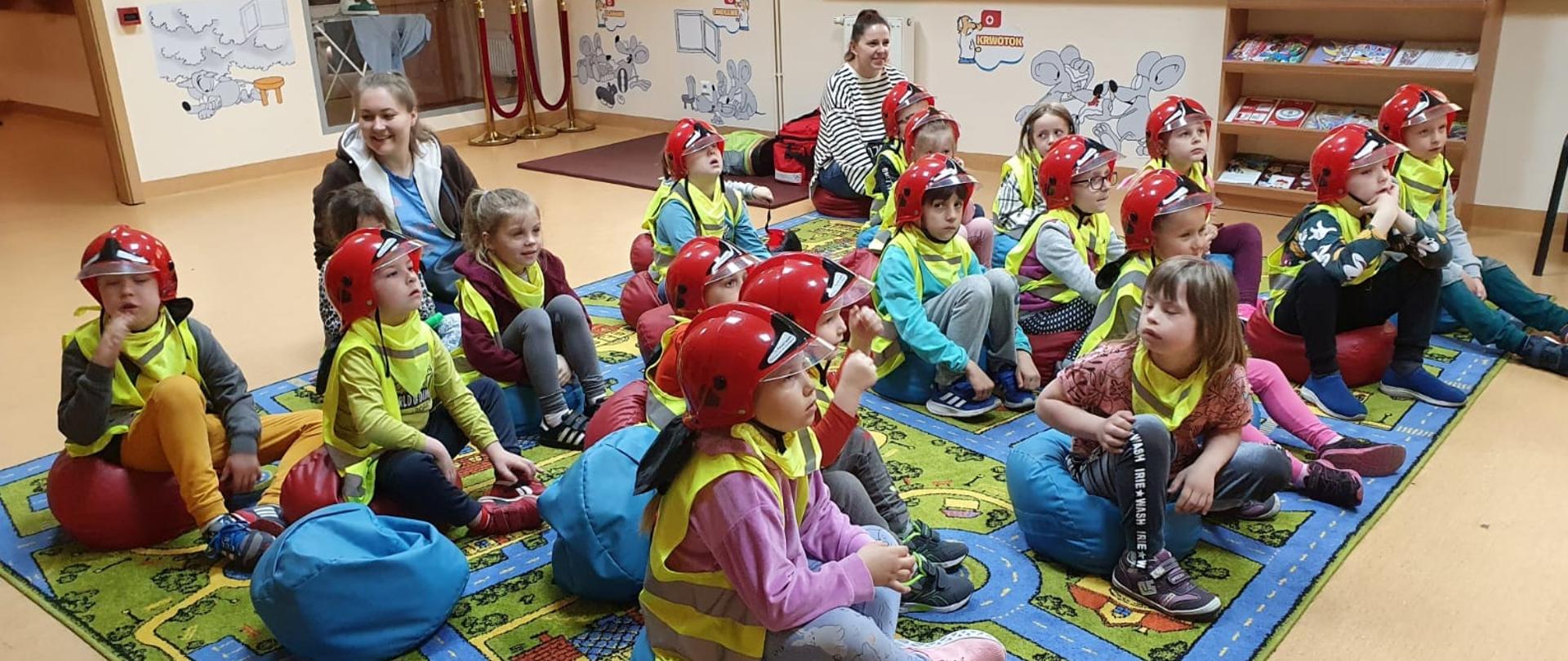 Dzieci w hełmach na głowach siedzą na kolorowych pufach. Pufy ustawione są na kolorowym dywanie. Za dziećmi siedzą dwie Panie. W tle widać rysunki na ścianach oraz szybę za którą stoi deska do prasowania a na niej żelazko.