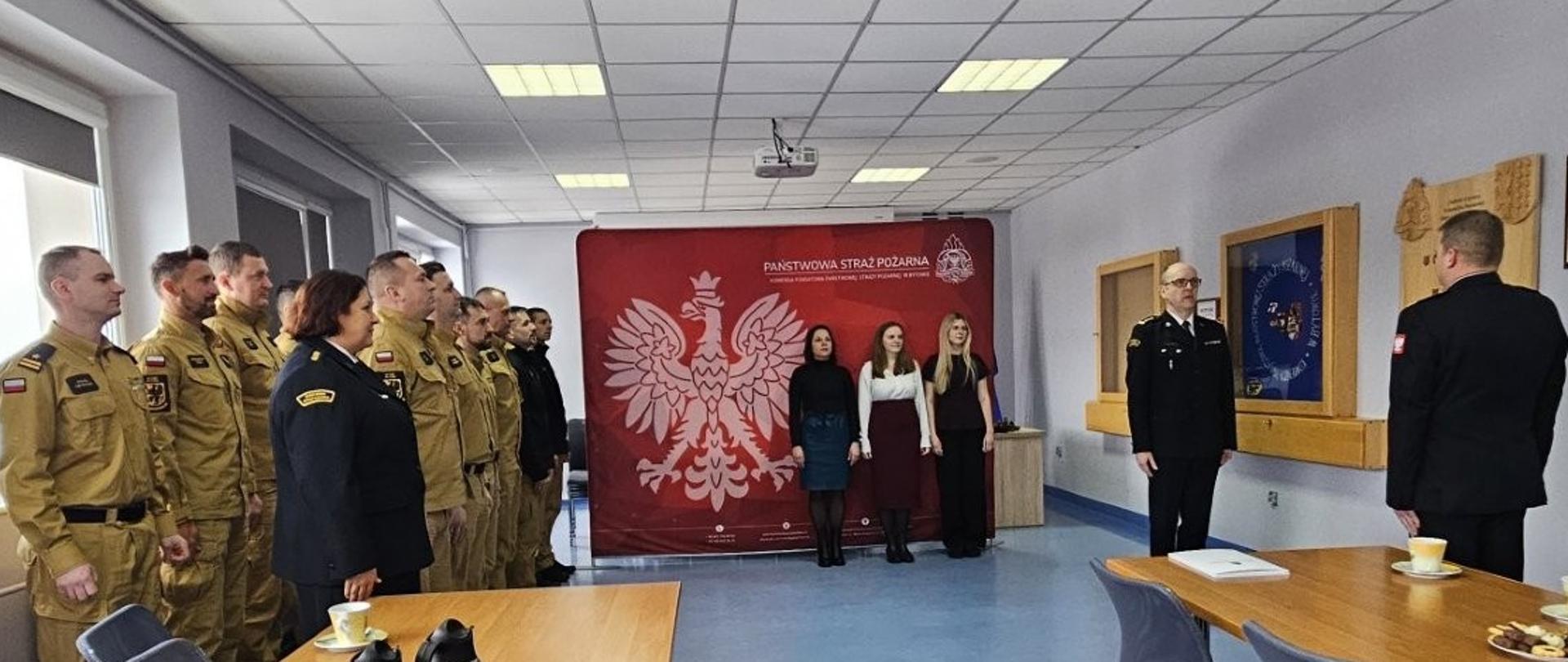 Funkcjonariusze PSP stoją z lewej strony zdjęcia pod oknem. Przed nimi zastępca komendanta powiatowego KP PSP w Bytowie składa meldunek komendantowi. W centralnej części zdjęcia stoją trzy kobiety na tle czerwonej ścianki z orłem oraz logiem PSP