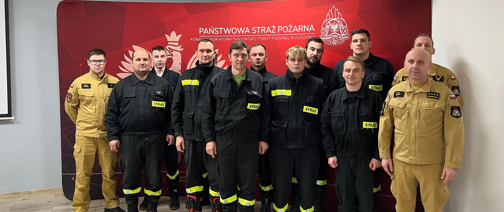 Grupa mężczyzn ubranych w mundury straży pożarnej stoi w dwu szeregu przed czerwonym banerem Państwowej Straży Pożarnej.
