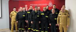 Grupa mężczyzn ubranych w mundury straży pożarnej stoi w dwu szeregu przed czerwonym banerem Państwowej Straży Pożarnej.