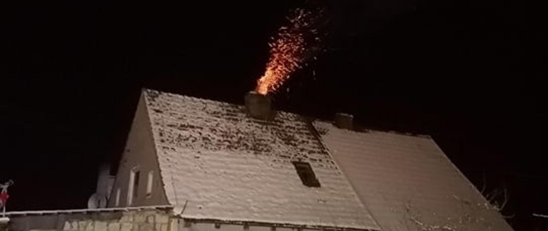 Zdjęcie przedstawia dom jednorodzinny, dach domu pokryty śniegiem. Z komina widać wydostający się ogień oraz iskry. Zdjęcie zrobione nocą. W tle widać ogródek zasypany śniegiem.