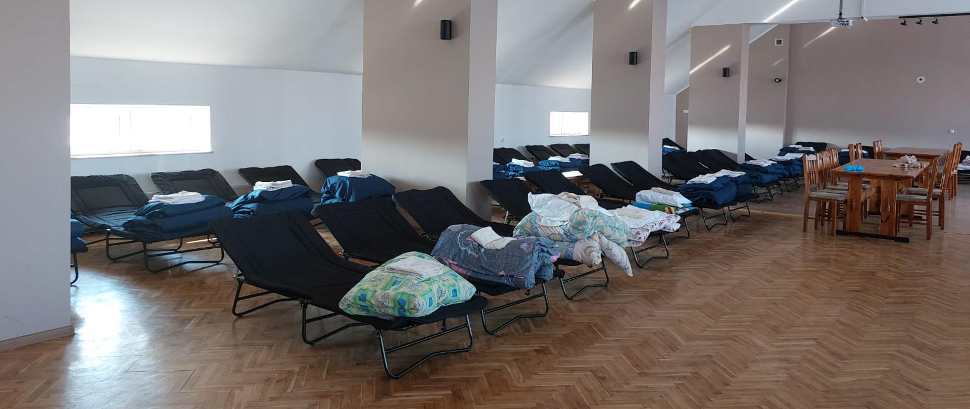 Zdjęcie wykonane w jednym z pomieszczeń wewnątrz strażnicy Ochotniczej Straży Pożarnej w Jastrzębi. Na zdjęciu widać poustawiane w dwóch rzędach, przygotowane do spania miejsca dla uchodźców z Ukrainy. Na łóżkach znajduje się kolorowa pościel. 