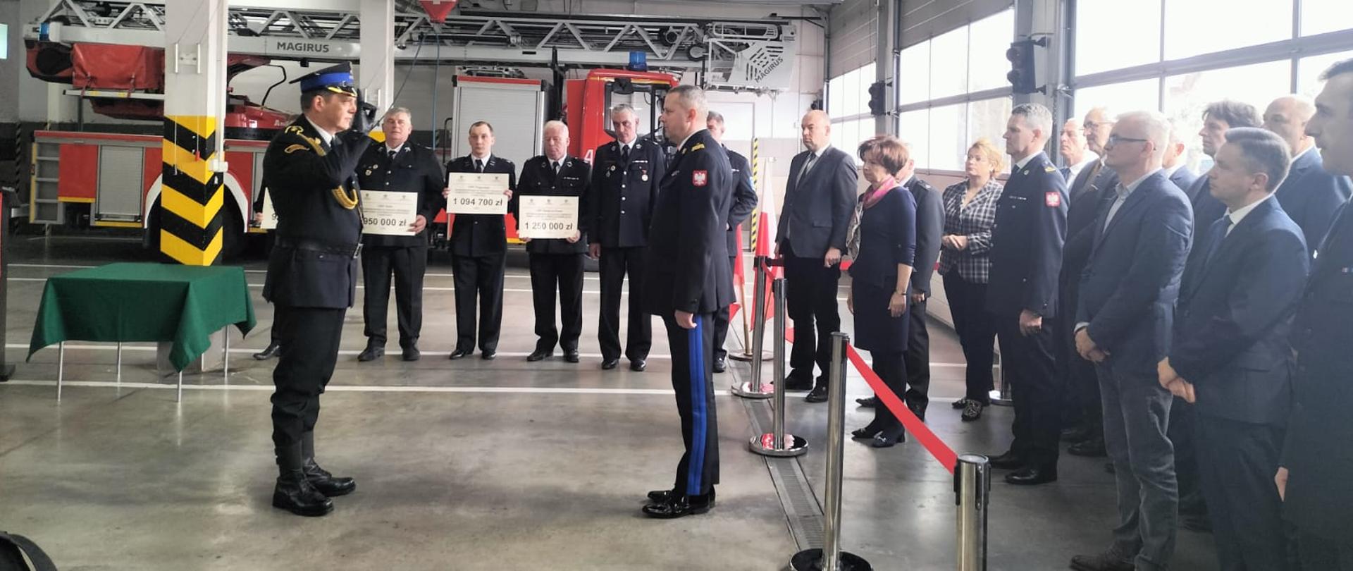 Zdjęcie przedstawia dowódcę uroczystości składającego meldunek Wielkopolskiego Komendantowi Wojewódzkiemu PSP. Dowódca oddaje honor poprzez salutowanie. W tle stoją druhowie OSP, którzy trzymają w rękach promesy. Z prawej strony stoją zaproszeni goście.
