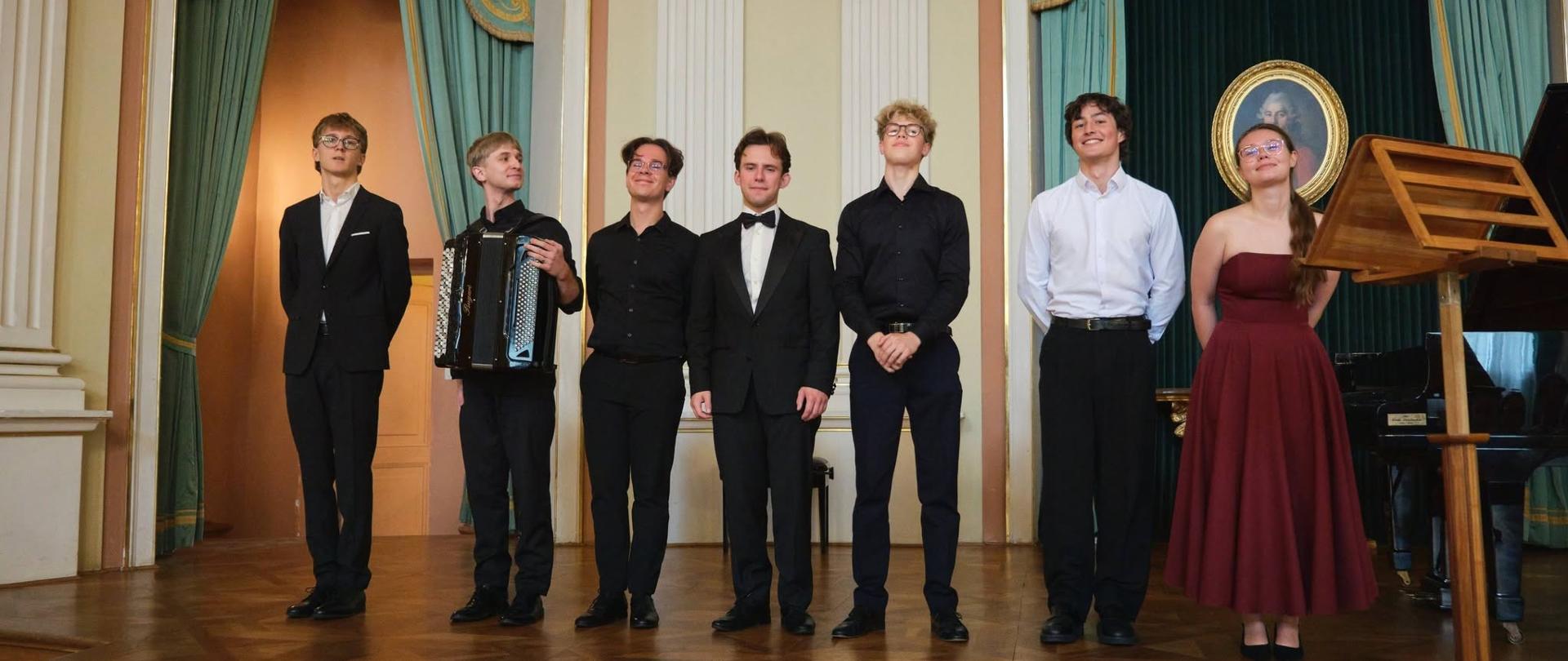 Zdjęcie przedstawia grupę siedmiorga młodych wykonawców (sześciu mężczyzn i kobietę) stojących w szeregu na scenie sali koncertowej o klasycznym wystroju. Wykonawcy: Wszyscy są ubrani w eleganckie, galowe stroje. Drugi od lewej mężczyzna trzyma trzymany na ramionach akordeon, a po prawej stronie stoi kobieta w długiej, bordowej sukni bez ramiączek. Wnętrze: Występ odbywa się w przestronnej sali z drewnianym parkietem, zdobionymi arkadami, zielonymi zasłonami oraz portretem w owalnej ramie zawieszonym w tle. Po prawej stronie na pierwszym planie widoczny jest drewniany pulpit na nuty, a za nim fragment fortepianu. 