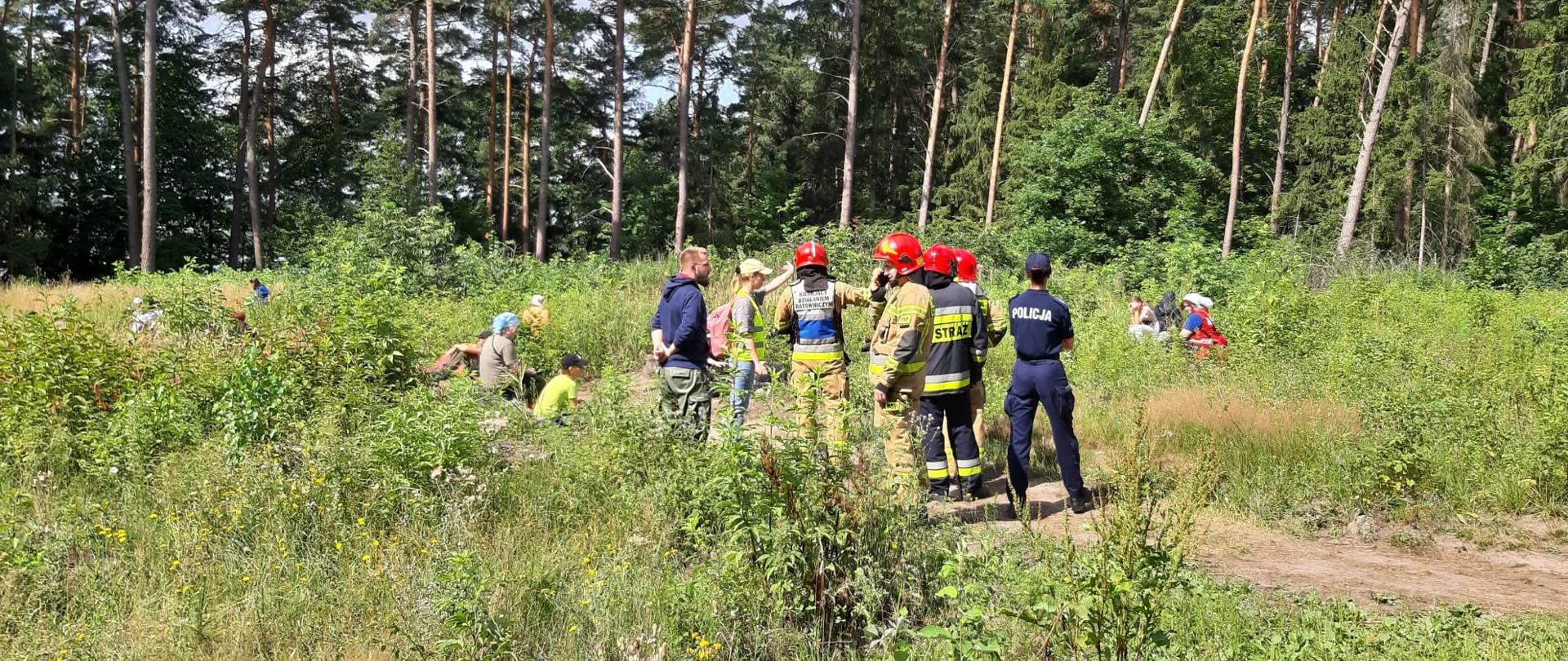 Na polanie znajdują się ewakuowane dzieci. Widać również strażaków wraz z policją. Strażaka zbiera informację od opiekunów ilu ma harcerzy. 