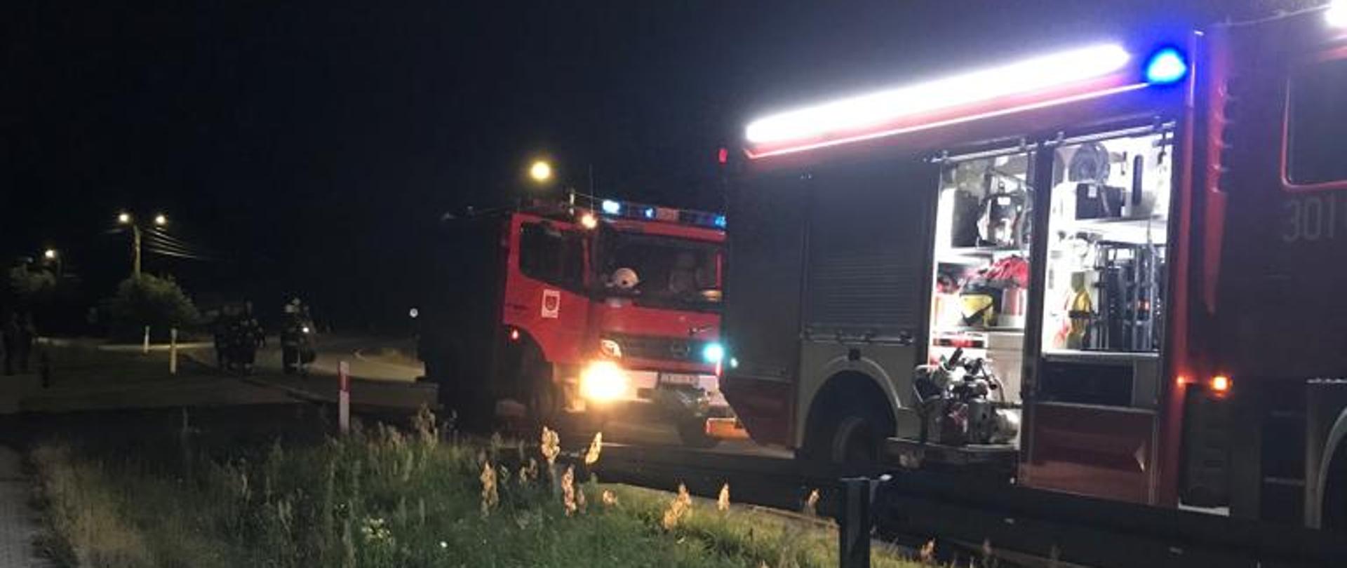 Zdjęcie przedstawia dwa samochody pożarnicze stojące na drodze, za pojazdami w pewnym oddaleniu grupa strażaków biorących udział w akcji.
