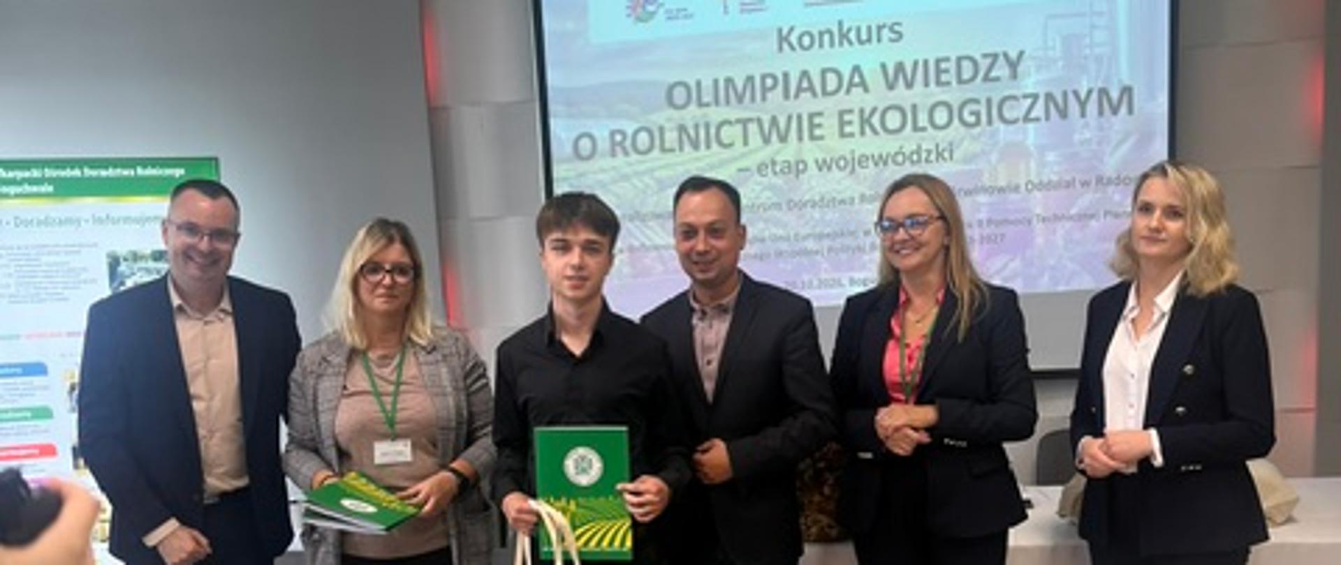 Grupowe zdjęcie uczestników i organizatorów Olimpiady Wiedzy o Rolnictwie Ekologicznym podczas uroczystości wręczenia nagród. Nagrodzeni uczestnicy trzymają dyplomy, upominki i materiały konkursowe