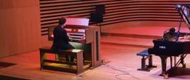 Mężczyzna siedząc tyłem do publiczności gra na organach, po prawej stronie stoi fortepian Fazioli.