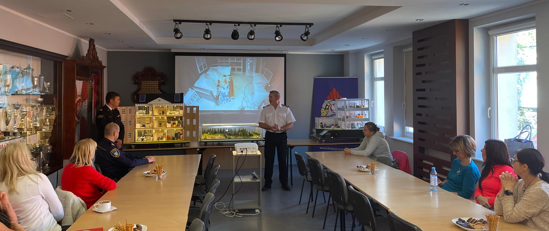 Świetlica komendy. Przy stołach ułożonych w dwóch rzędach siedzą uczestnicy. Z tyłu sali ustawione na stołach mobilne symulatory zagrożeń przy kturych stoi dwóch strażaków. komendant wita uczestników