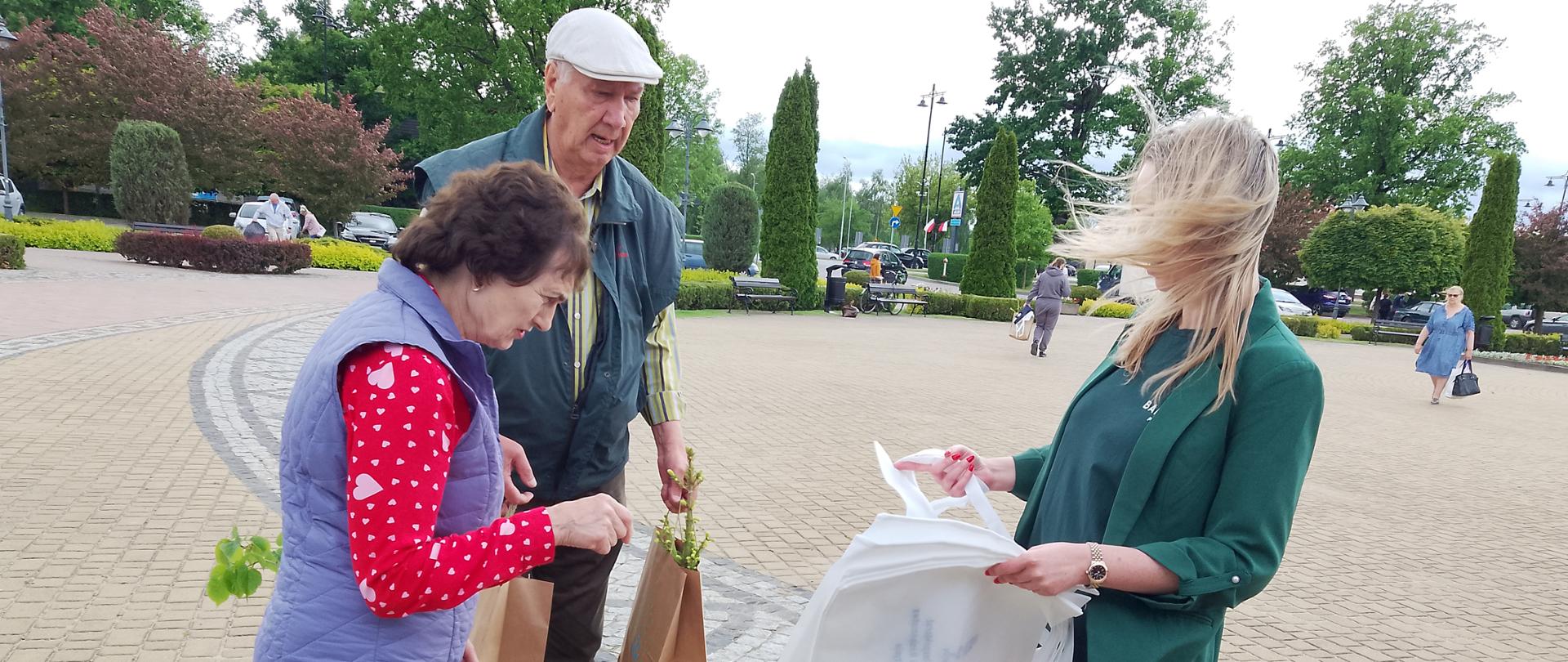 Na placu miejskim kobieta stoi naprzeciwko starszego małżeństwa, rozmawiają, trzymają w rękach torby z roślinami