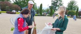 Na placu miejskim kobieta stoi naprzeciwko starszego małżeństwa, rozmawiają, trzymają w rękach torby z roślinami