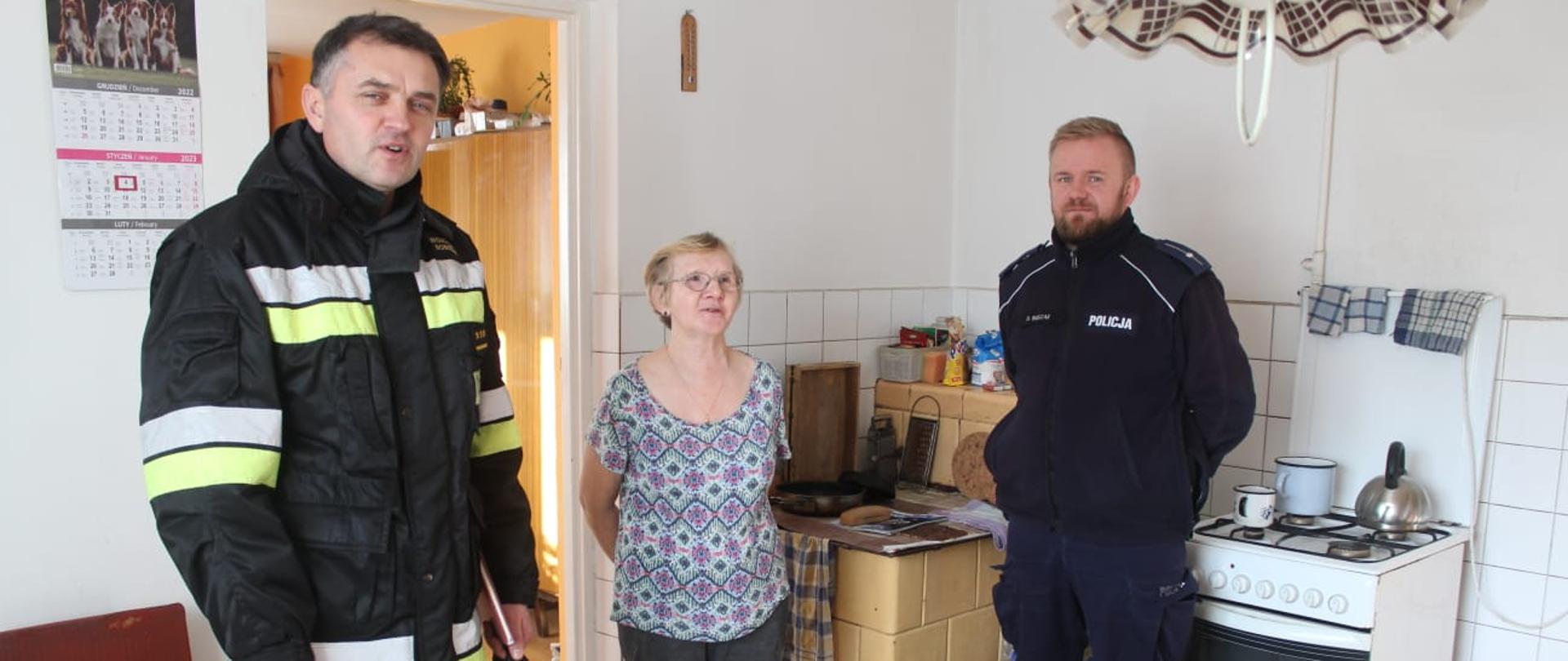 Kolorowa fotografia wykonana w słoneczny dzień w budynku mieszkalnym. Przedstawia na pierwszym planie strażaka w ubraniu dowódczo sztabowym i policjanta, natomiast między nimi właścicielka budynku. Fotografia wykonana w pomieszczeniu kuchni, za osobami przedstawionymi wyżej znajdują się urządzenia grzewcze (kuchnia gazowa i kaflowa). 