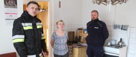 Kolorowa fotografia wykonana w słoneczny dzień w budynku mieszkalnym. Przedstawia na pierwszym planie strażaka w ubraniu dowódczo sztabowym i policjanta, natomiast między nimi właścicielka budynku. Fotografia wykonana w pomieszczeniu kuchni, za osobami przedstawionymi wyżej znajdują się urządzenia grzewcze (kuchnia gazowa i kaflowa). 
