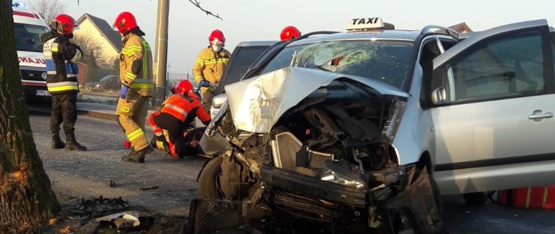 Zdjęcie przedstawia samochód osobowy, który w wyniku zderzenia całkowitemu zniszczeniu uległ przód samochodu a przednia szybka została zbita. Za samochodem są strażacy zabezpieczający miejsce zdarzenia oraz pomagający zespołowi ratownictwa medyczne w działaniach ratowniczych. W tle po lewej stronie stoi karetka pogotowia ratunkowego. 