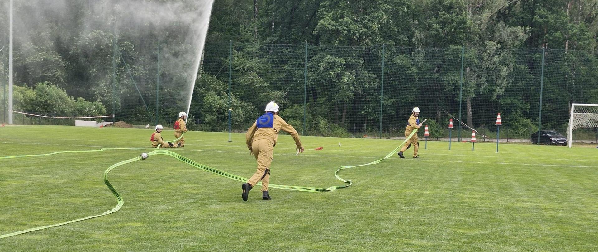 Zdjęcie dokumentuje kluczowy moment konkurencji bojowej, rozgrywanej na zielonej murawie stadionu sportowego. W akcji bierze udział co najmniej czworo strażaków ochotników.
Centralny plan: Na pierwszym planie, tyłem do obiektywu, widoczna jest strażaczka w piaskowym mundurze koszarowym i hełmie, która biegnie w stronę linii wodnej, trzymając w rękach rozwinięty wąż strażacki.
Lewa strona: W głębi, przy rozdzielaczu, widać dwóch innych strażaków. Jeden z nich, klęcząc przy prądownicy, kieruje silny strumień wody na tarczę celowniczą (niewidoczną w kadrze, ale sugerowaną przez kierunek strumienia i rozprysk wody).
Prawa strona: W głębi, na wysokości tyczek wyznaczających tor, stoi kolejny strażak w hełmie, najprawdopodobniej asekurujący linię wężową lub czekający na swoją kolej.
Tło: Zbiórka odbywa się na tle wysokiego ogrodzenia, za którym znajduje się gęsty, zielony las. Na murawie widoczne są tyczki sygnalizacyjne, co potwierdza, że jest to wyznaczony tor zawodów. Widać również fragment bramki piłkarskiej na skraju boiska.
Atmosfera: Ujęcie doskonale oddaje dynamikę i skupienie towarzyszące zawodom OSP, gdzie liczy się każda sekunda i precyzja podania wody.