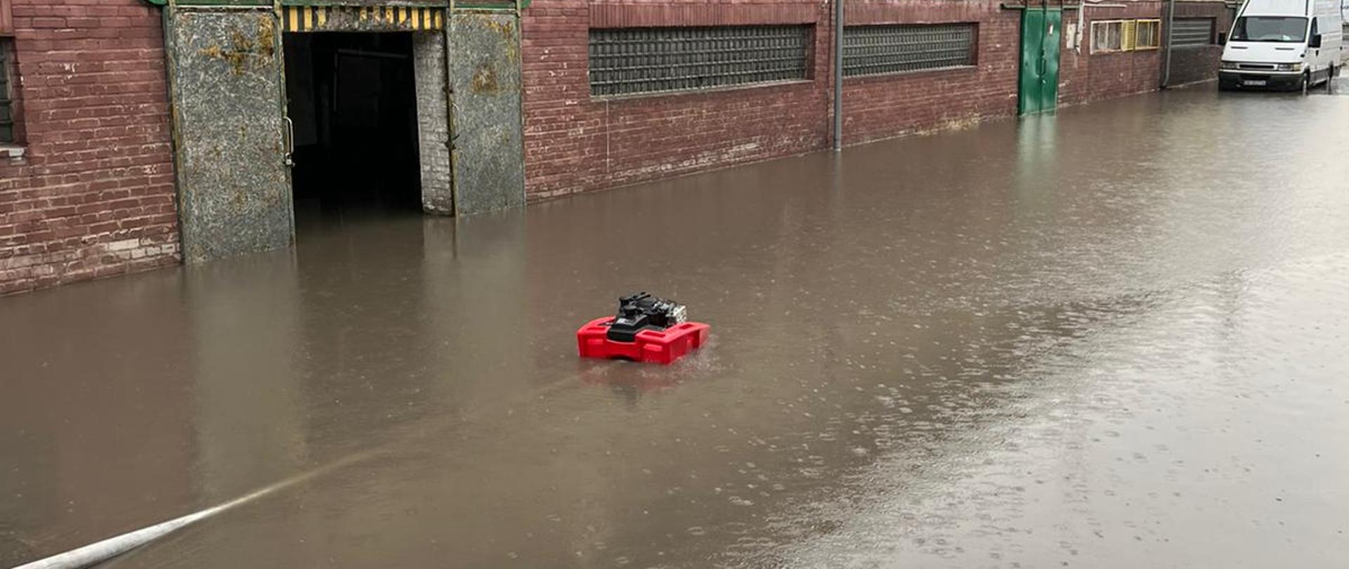 Pompa pływająca pompuje wodę zgromadzoną na placu oraz w obiekcie magazynowo-produkcyjnym. W tle widoczny samochód dostawczy koloru białego.