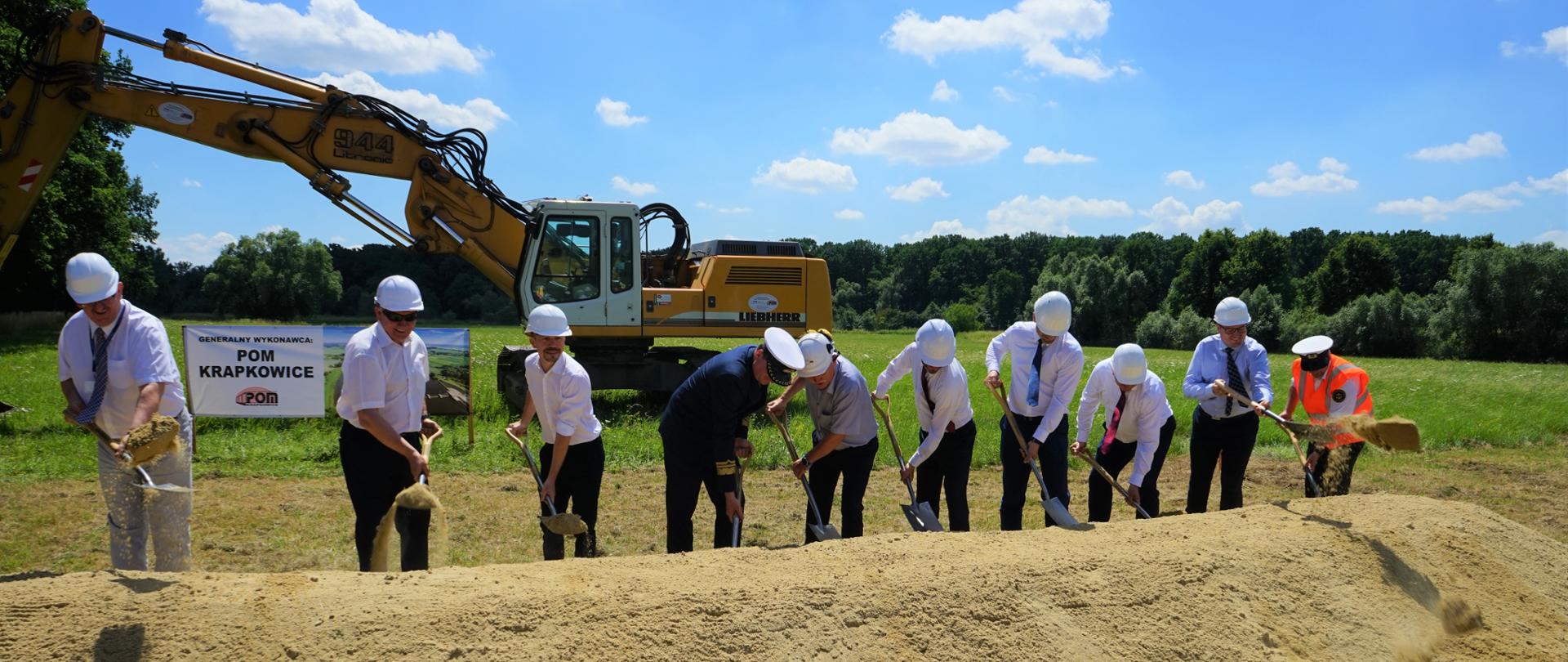 Na zdjęciu 10 mężczyzn - większość w mundurach kapitana statku. Mężczyźni stoją na tle koparki oraz łąki. Panowie wbijają łopaty pod inwestycję - na znak startu prac. 