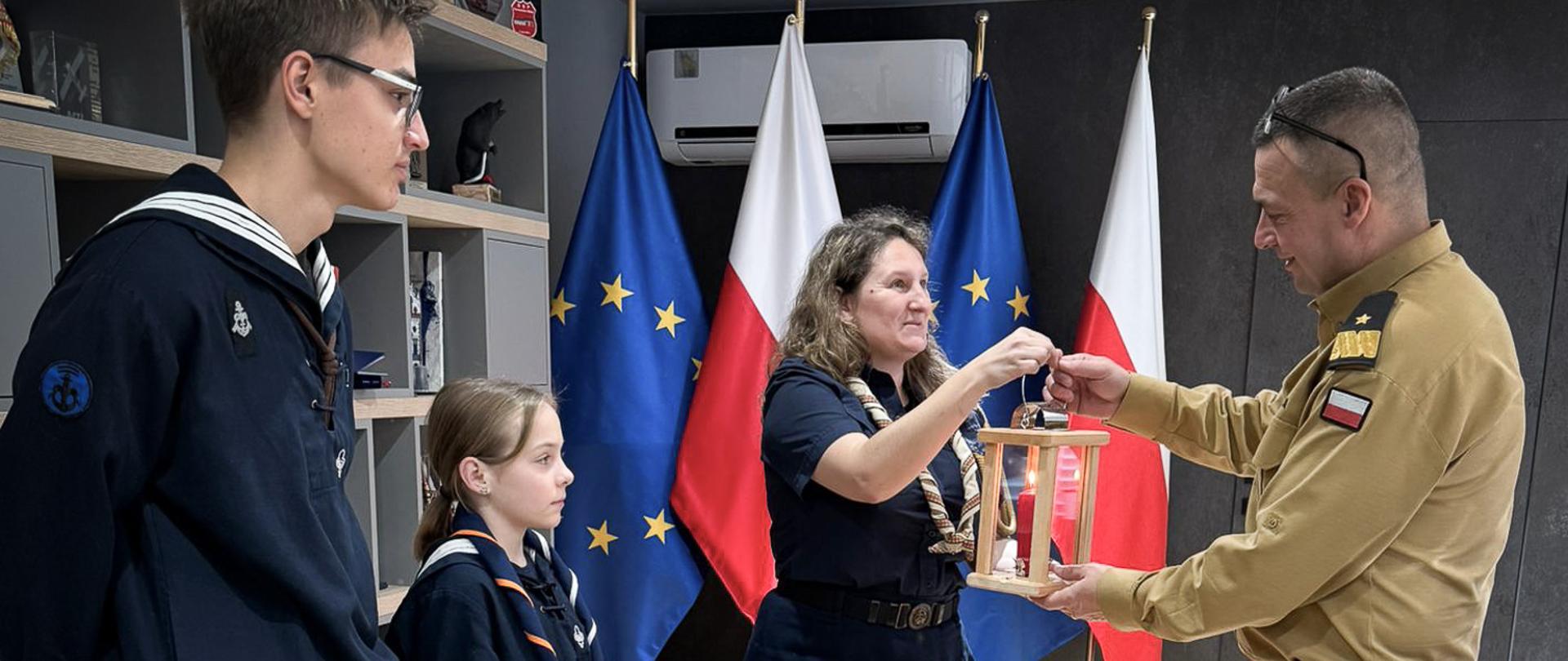 Zdjęcie zrobione wewnątrz. Harcerz przekazuje świecę betlejemskiego światła pokoju generałowi Państwowej Straży Pożarnej. W obok stoją harcerze. Za nimi ustawione są flagi.