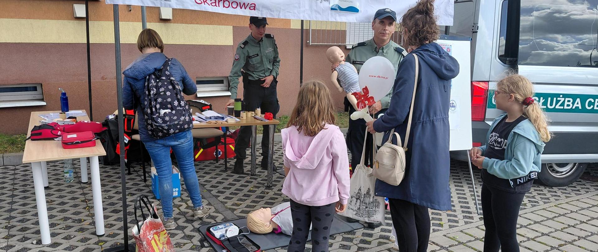 2 funkcjonariuszy pokazuje na fantomach pierwszą pomoc, z boku 2 kobiety i 2 dzieci
