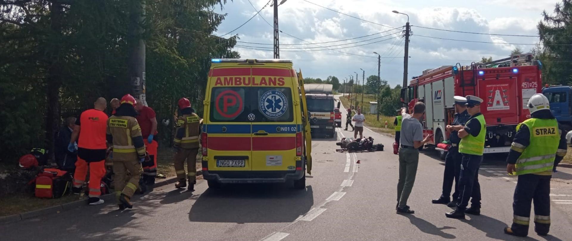 Zdjęcie przedstawia motocykl po zderzeniu z pojazdem ciężarowym leżący na środku drogi wojewódzkiej nr 650. Po lewej stronie widać karetkę pogotowia oraz strażakaków i ratowników medycznych, którzy udzielają pomocy medycznej poszkodowanemu motocykliście. Natomiast po prawej stronie można zobaczyć pojazd pożarniczy oraz policjantów i strażaków zabezpieczających miejsce zdarzenia