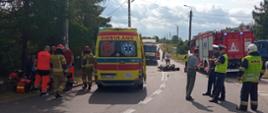 Zdjęcie przedstawia motocykl po zderzeniu z pojazdem ciężarowym leżący na środku drogi wojewódzkiej nr 650. Po lewej stronie widać karetkę pogotowia oraz strażakaków i ratowników medycznych, którzy udzielają pomocy medycznej poszkodowanemu motocykliście. Natomiast po prawej stronie można zobaczyć pojazd pożarniczy oraz policjantów i strażaków zabezpieczających miejsce zdarzenia