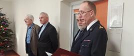 Zdjęcie przedstawia Komendanta Powiatowego PSP w Świdwinie podczas czytania życzeń świątecznych, obok niego znajduję się wice starosta, ksiądz oraz najstarszy z obecnych byłych Komendantów tutejszej jednostki. panowie znajdują się na świetlicy na której znajduję się udekorowana choinka.