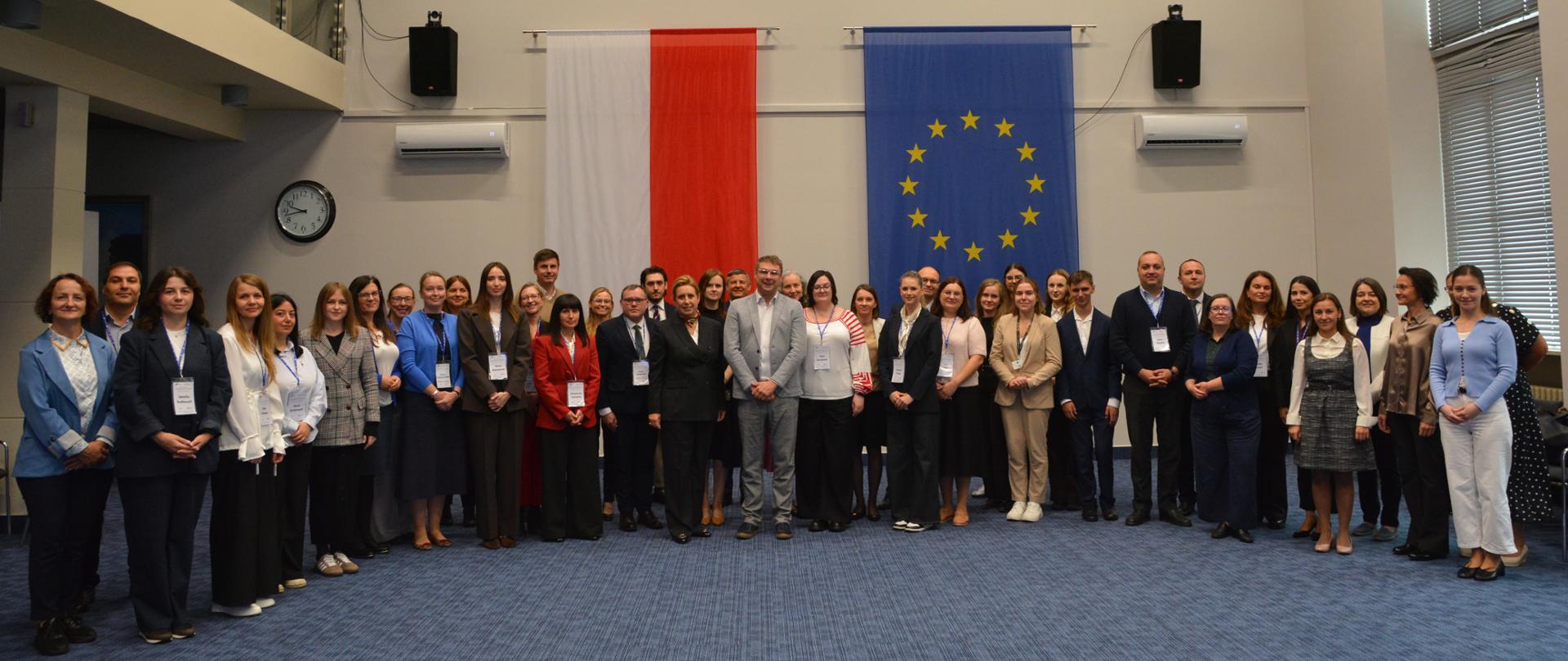 Duża grupa kobiet i mężczyzn stoi w półkolu w dużej sali. Nad nimi wiszą: godło Polski oraz flaga polska i flaga Unii Europejskiej 