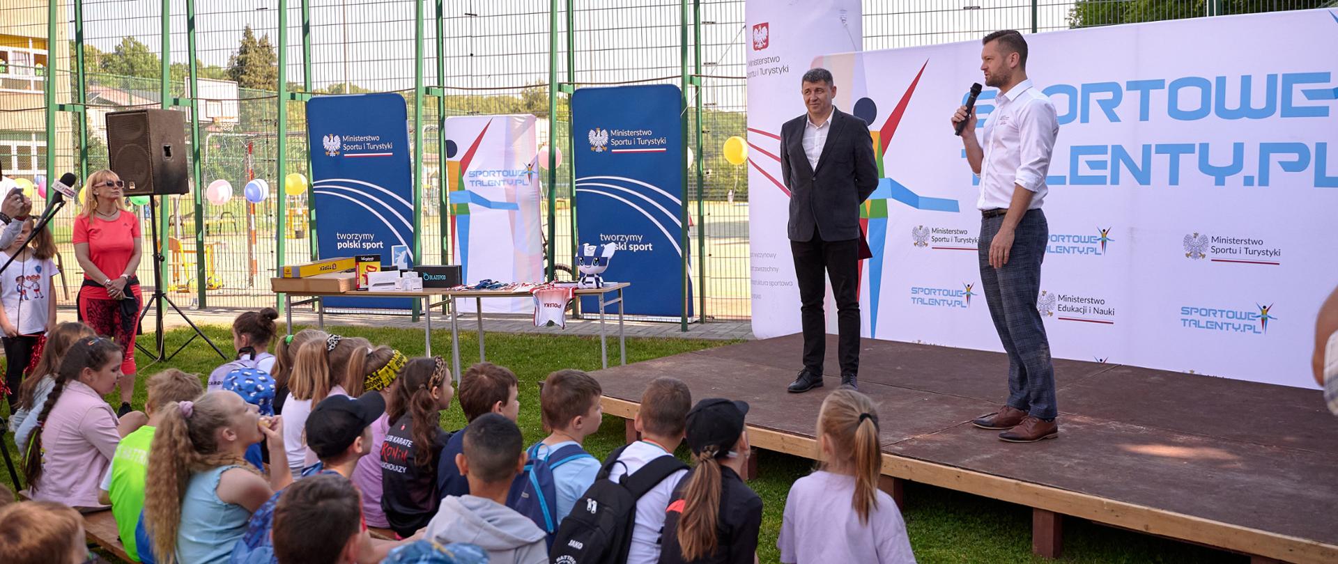 Minister Kamil Bortniczuk przemawia do dzieci, stojąc na scenie. Dzieci siedzą na ławkach. W tle napis na bannerze - Sportowe Talenty
