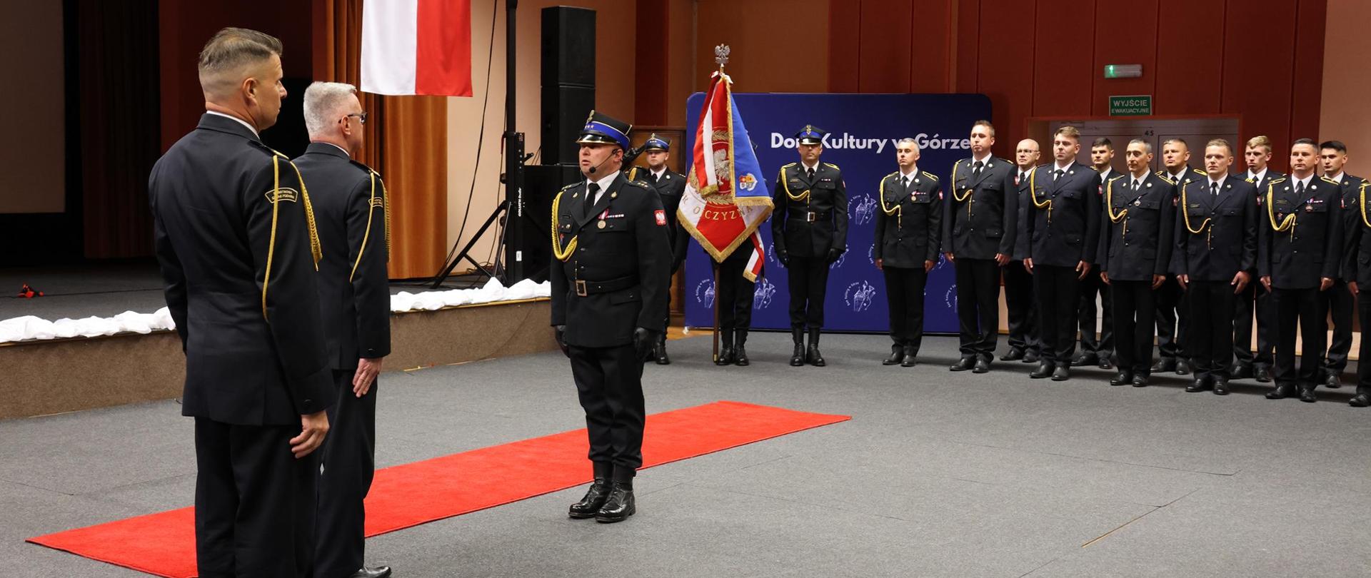 Wnętrze sali widowiskowej. Na środku sali, na czerwonym dywanie, stoi grupa umundurowanych funkcjonariuszy straży pożarnej w galowych strojach. Jeden z nich, stojący przodem do dwóch pozostałych, składa meldunek. Za nim znajduje się osoba trzymająca sztandar jednostki. Pozostali funkcjonariusze stoją w szeregu po prawej stronie, zachowując postawę wyprostowaną. W tle widać zawieszoną polską flagę Polski oraz oświetlenie sceniczne. Zdjęcie ma charakter uroczystej ceremonii.