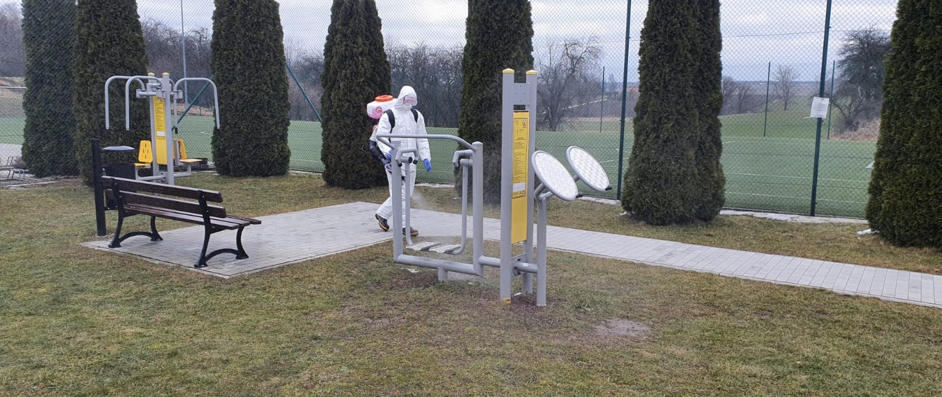 Strażak w ubraniu ochronnym dezynfekuje przy pomocy opryskiwacza spalinowego siłownię plenerową.