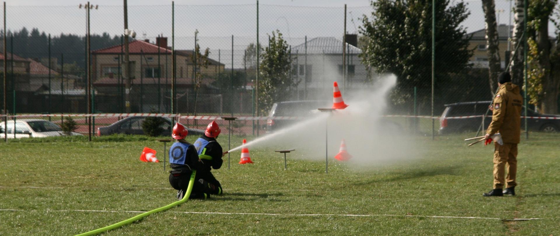 Zdjęcie przedstawia moment przebiegu konkurencji ćwiczenie bojowe przez męską drużynę pożarniczą podczas VIII Powiatowych Zawodów Sportowo-Pożarniczych Ochotniczych Straży Pożarnych powiatu gołdapskiego (zrzucenie pachołków z podstaw prądami wody)