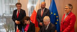Przy nakrytym niebieskim materiałem stoliku siedzi kobieta w czarnej marynarce i mężczyzna w czarnym garniturze, podpisują dokumenty, nad nimi stoi z prawej wiceminister Mrówczyńska w czerwonym garniturze i dwóch mężczyzn w garniturach, za nimi pod ścianą flagi Polski i UE.